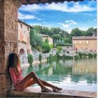 Two women by calm pond near red-brick buildings and greenery under blue sky.