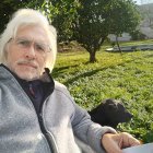 Elderly man with white beard and sunglasses beside a black dog, tram and greenery in background