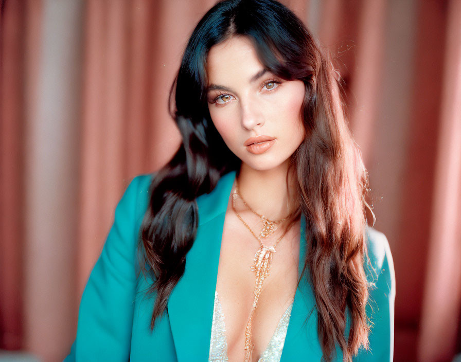 Woman with long brown hair in teal blazer and layered necklaces on blurred reddish background