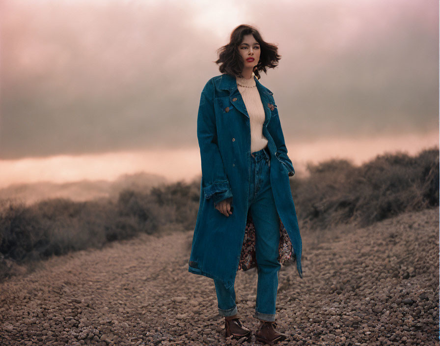 Woman in denim coat and jeans on gravel path with overcast skies