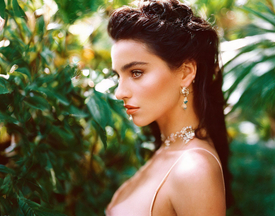 Dark-haired woman in updo with earrings and necklace poses in lush green foliage