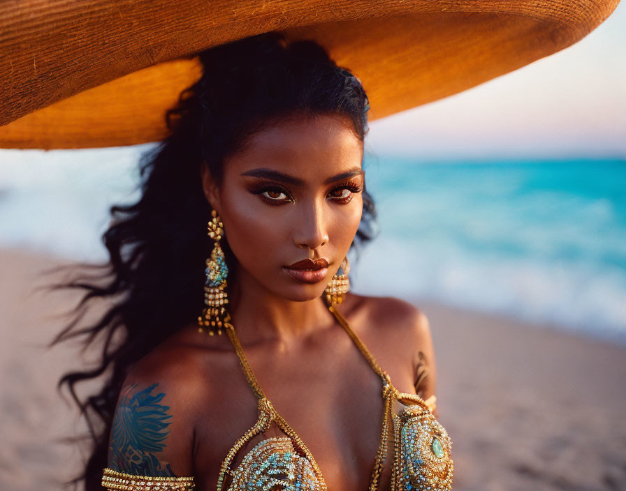 Woman with bold makeup and large hat on beach at dusk with waves and sand, adorned with intricate jewelry