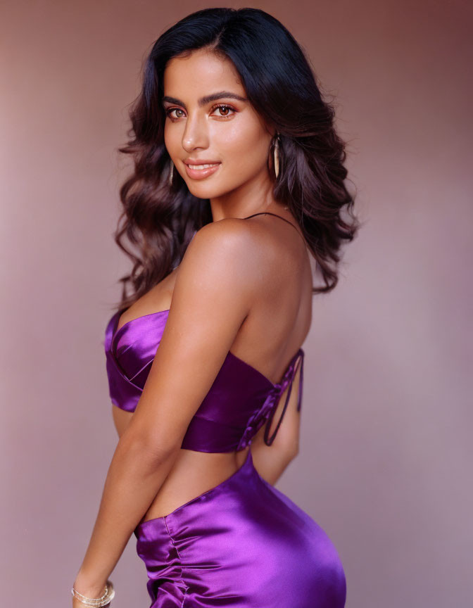 Woman in satin purple dress with wavy hair and hoop earrings on pink background