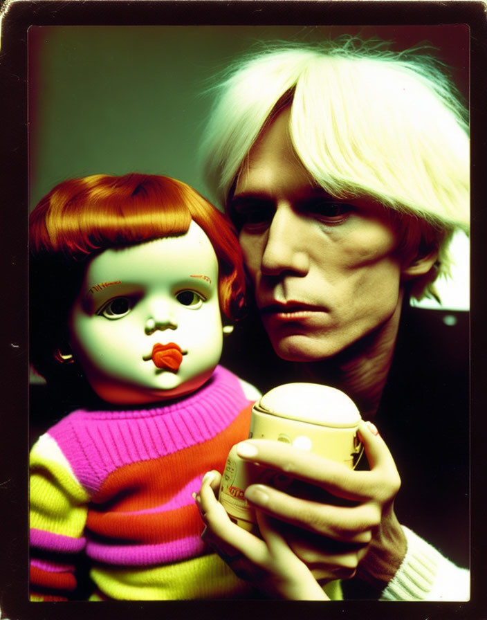 Pale person with white hair holding toy doll and cup on dark background