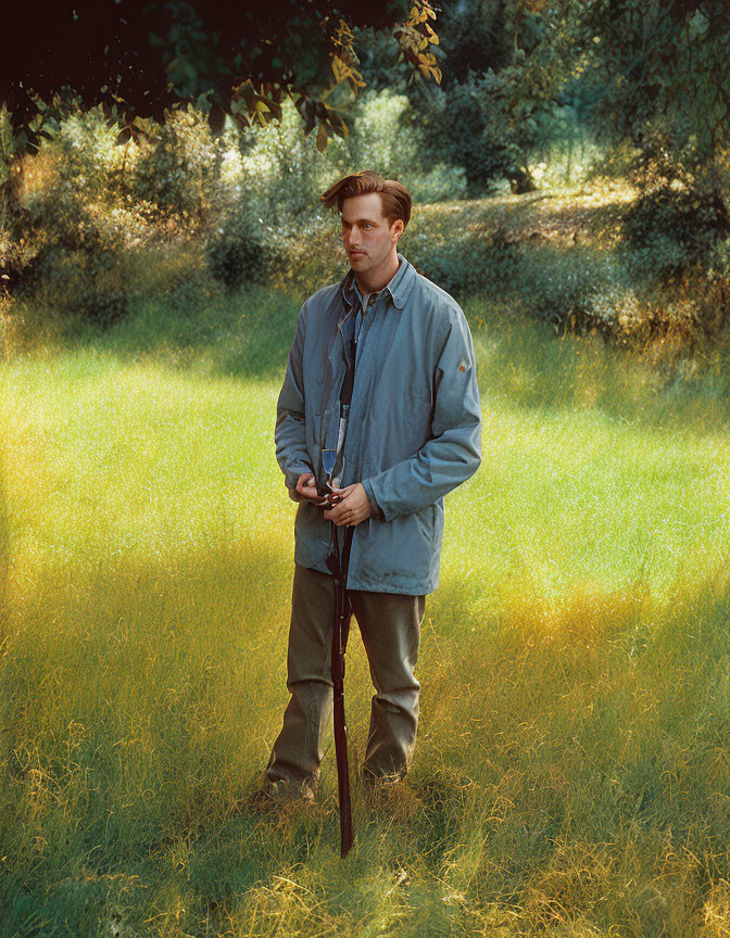 Man in sunlit field with trees, holding walking stick, wearing casual attire, looking contemplative.