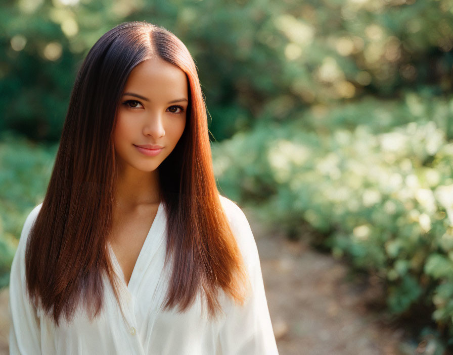 Woman with long brown hair in white blouse outdoors with greenery.