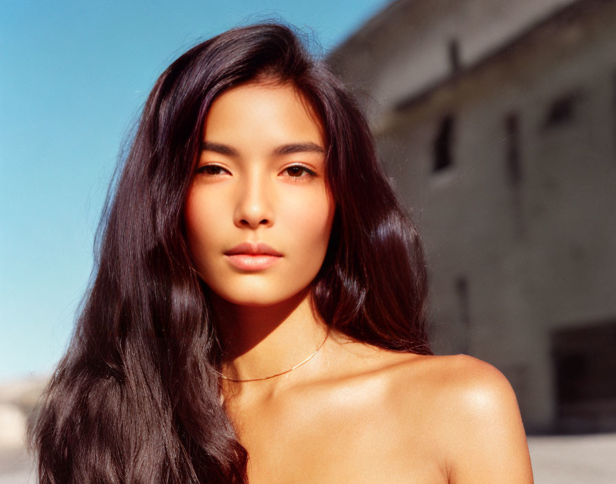 Dark-haired woman with bare shoulders in urban setting under warm sunlight.