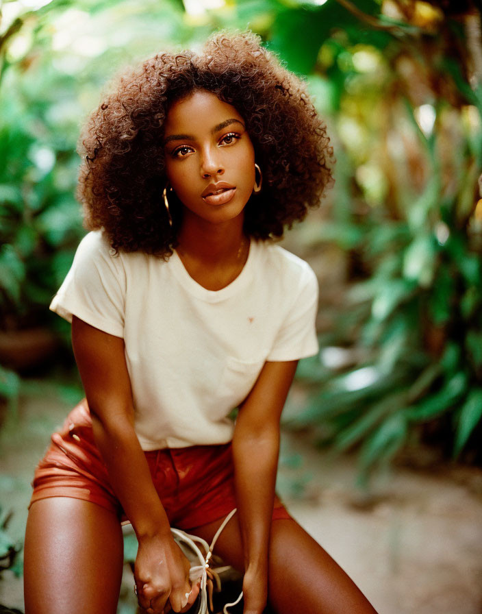 Curly-haired woman in cream top and terracotta shorts poses in garden.