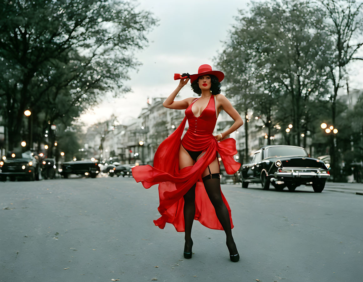 Woman in red dress and hat poses on tree-lined street with vintage cars