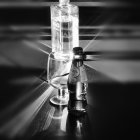 Monochrome photo of perfume bottle, vial, and glass on reflective surface