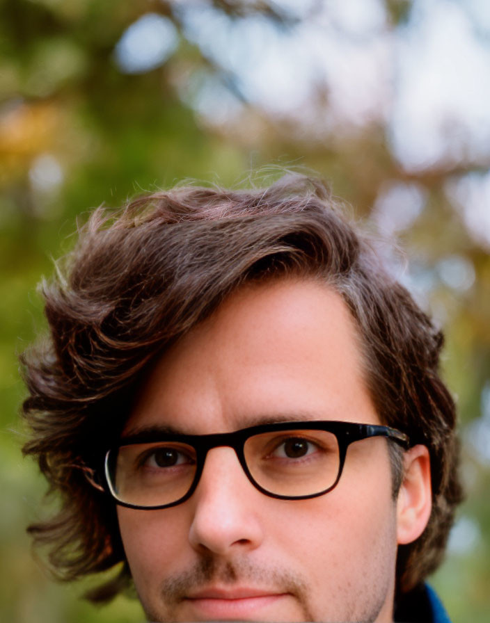 Portrait of person with glasses and tousled brown hair against autumnal backdrop.