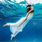 Woman in tribal headdress floats underwater in white gown amid surreal wave environment