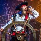 Female pirate with red hair steering ship's wheel in flamboyant attire against ghostly ship backdrop.