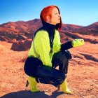 Red-haired person in neon green top crouching in desert with futuristic backdrop