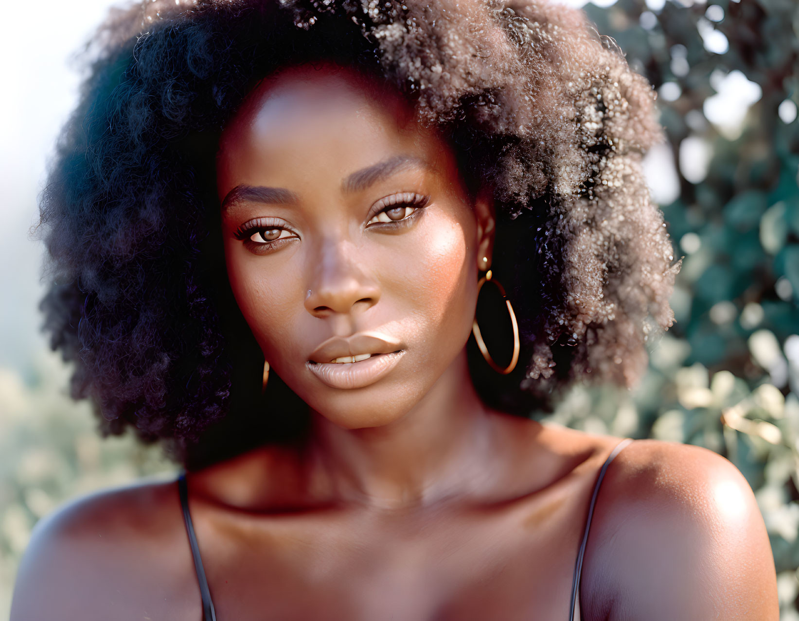 Woman with radiant skin and curly hair in hoop earrings and strap top on soft green backdrop