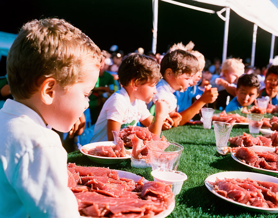 Kids in outdoor watermelon eating contest on sunny day.