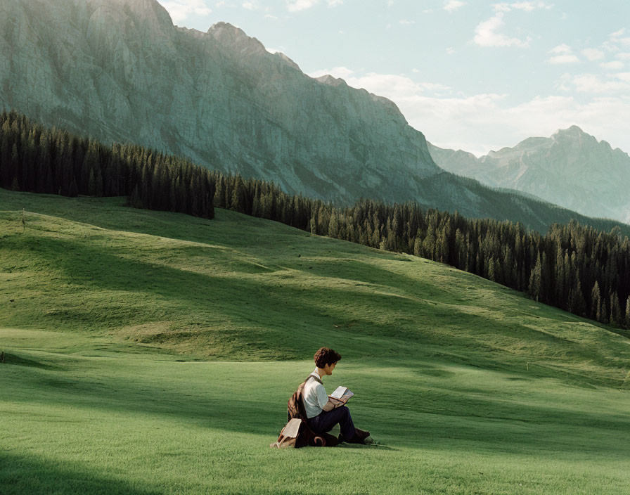 Person reading book on grassy hill with mountain backdrop