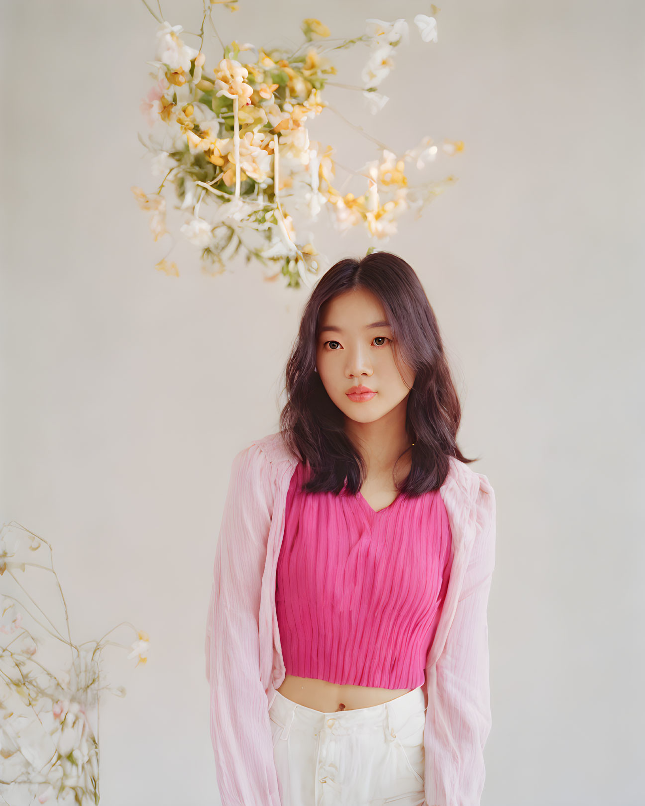 Woman in pink top and white pants under hanging white flowers on neutral background.