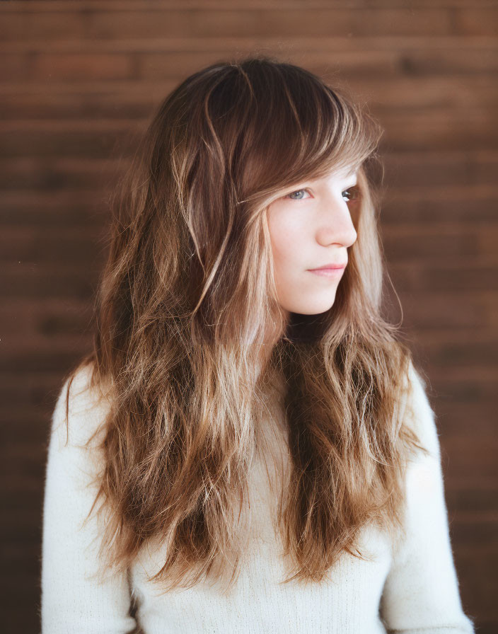 Young woman with long wavy brown hair in white sweater against wooden background