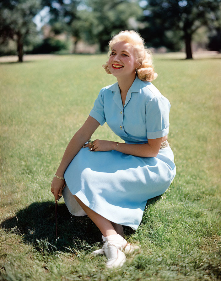 Smiling woman in blue dress sitting on grass with trees in background