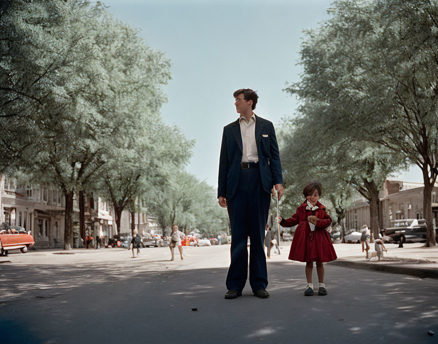 Man in suit with girl in red coat on tree-lined street with vintage cars.