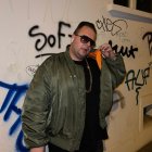 Fashionable person in black bomber jacket with gold accessories posing with spray paint gun against blue splattered wall