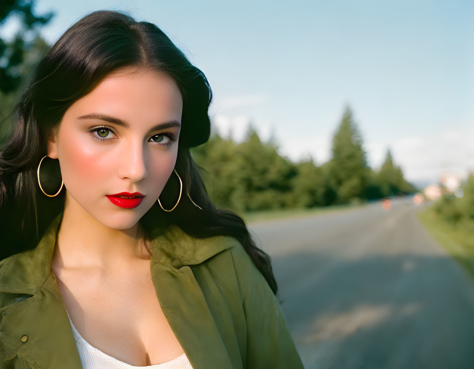 Young woman in red lipstick and hoop earrings, green jacket, roadside setting.