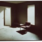 Empty dimly lit room with chair, stool, open door, two windows, and scattered debris.