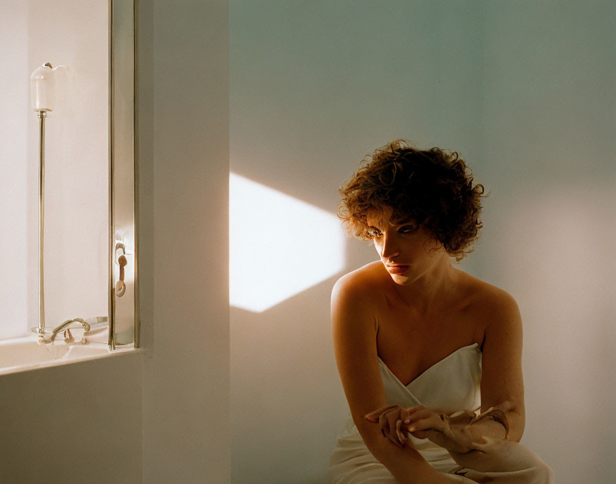 Curly-Haired Person Contemplating by Sunlit Window and Bathtub