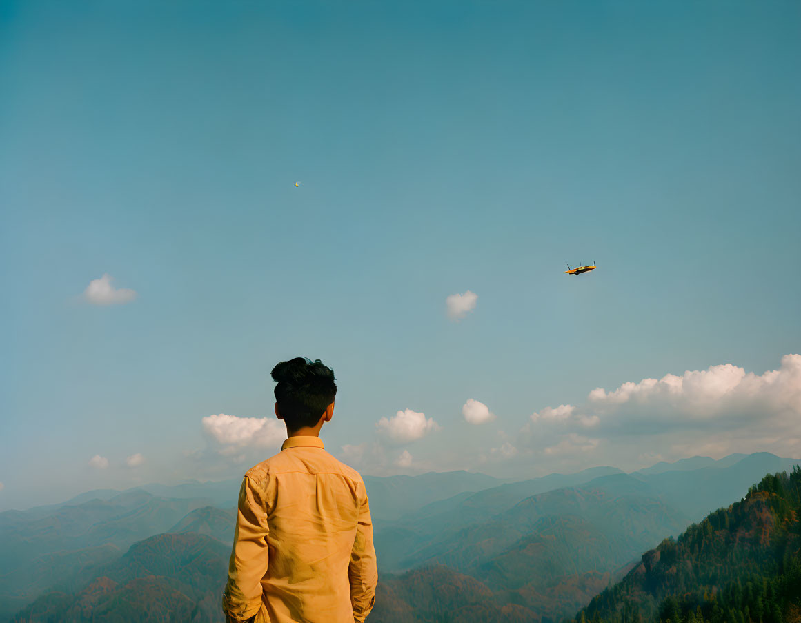 Person in yellow shirt admires mountain range and plane in blue sky