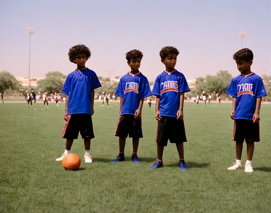 Four children in blue sports jerseys and black shorts on grassy field with soccer ball