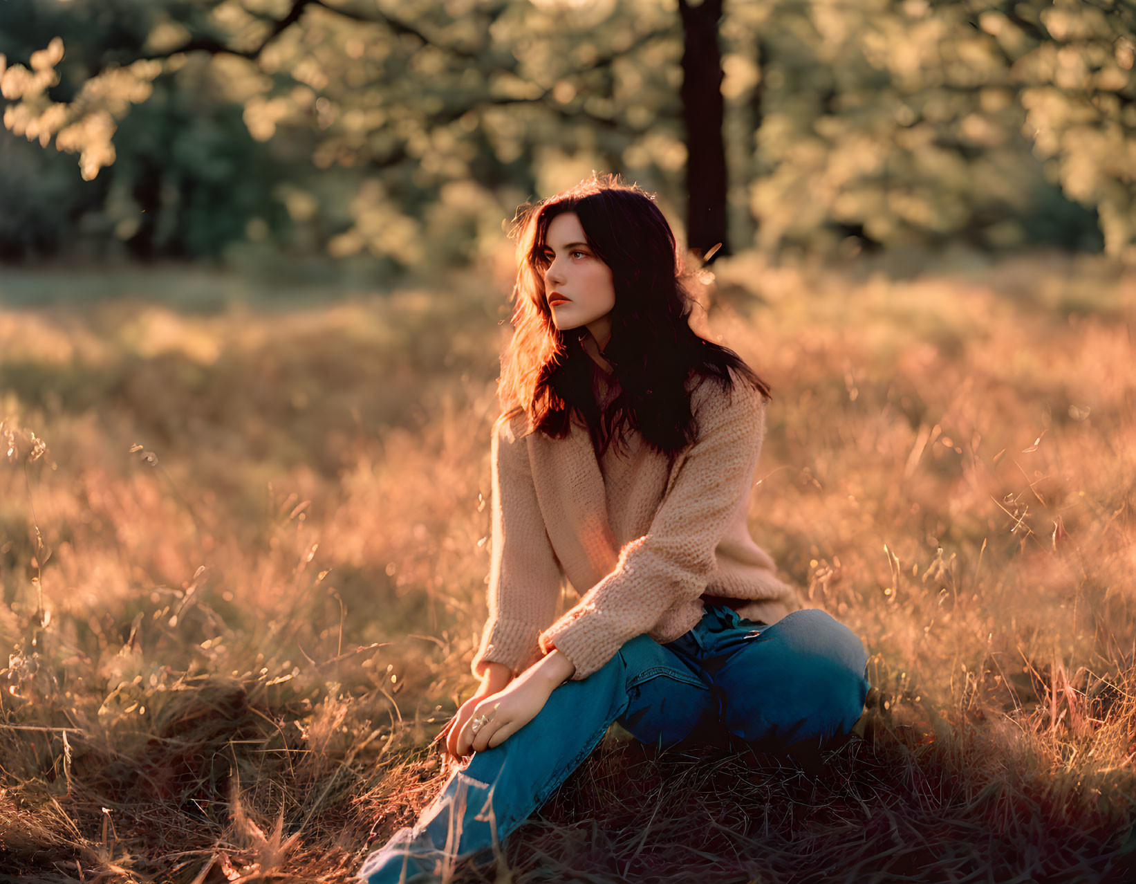 Woman in beige sweater and jeans sitting in sunlit field surrounded by trees