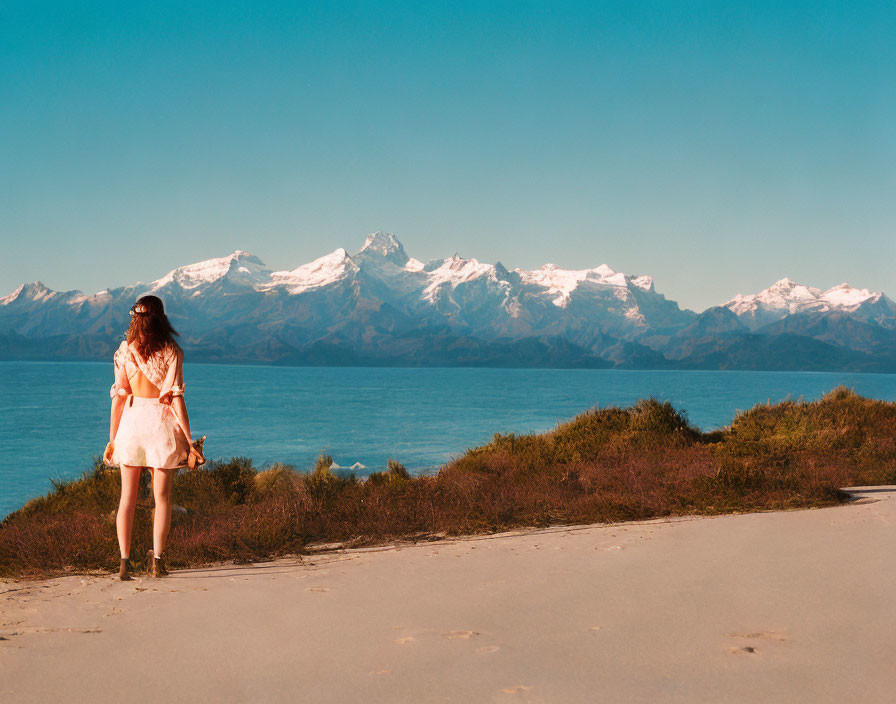 Woman in light dress gazes at tranquil lake and snow-capped mountains