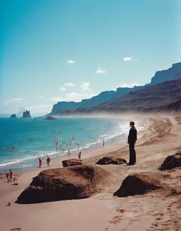 Person on sandy beach with ocean, people, and cliffs under clear blue sky