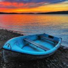 Turquoise boat painting on pebbled shore with sailboats under fiery sunset.