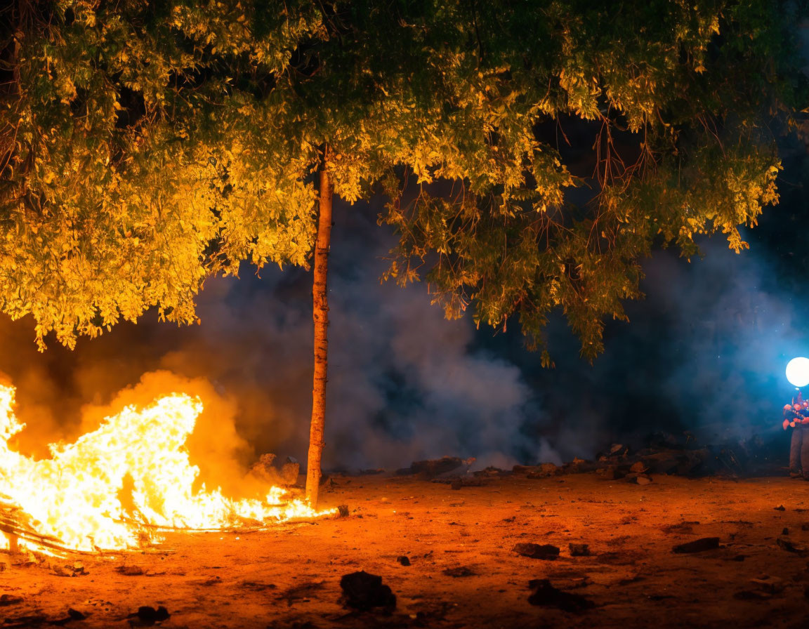 Nighttime bonfire under tree with figures and drifting smoke.