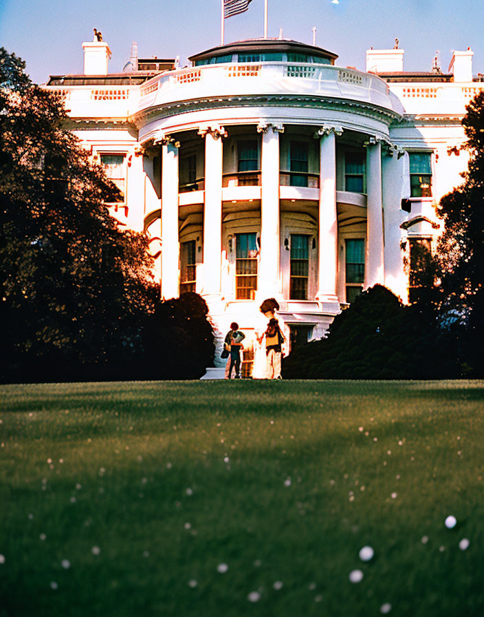 Sunset view of two individuals walking in front of illuminated White House façade