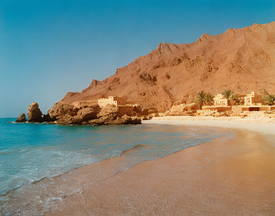 Golden sand beach with clear blue waters, mountain backdrop, and traditional buildings.