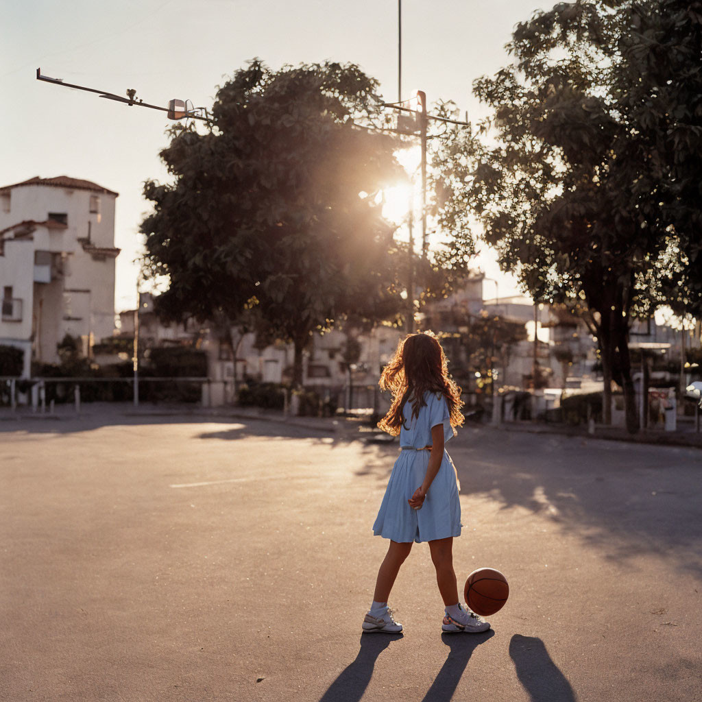 Girl in blue dress plays basketball on outdoor court at sunset