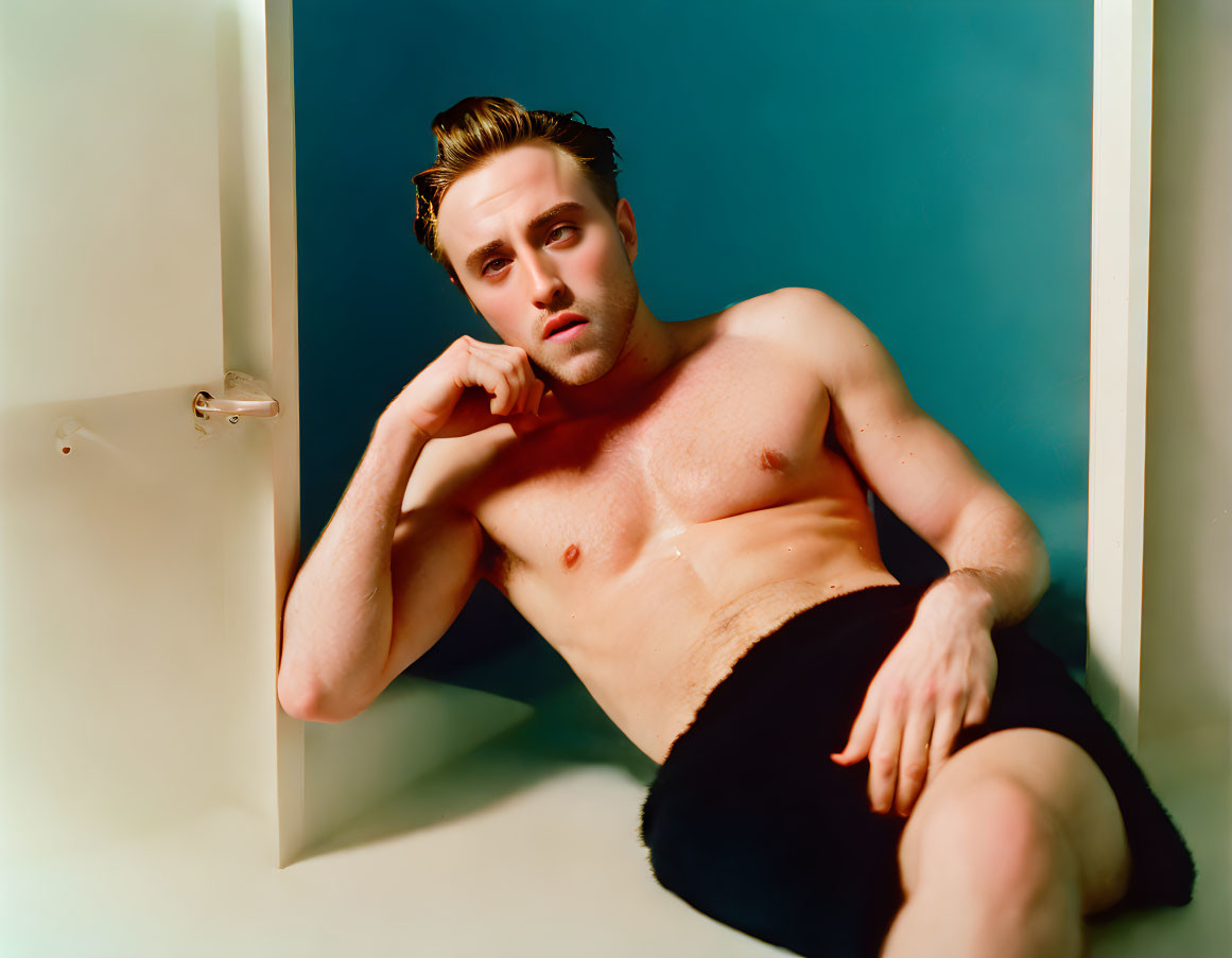 Shirtless man with slicked-back hair sitting by bathtub wrapped in towel