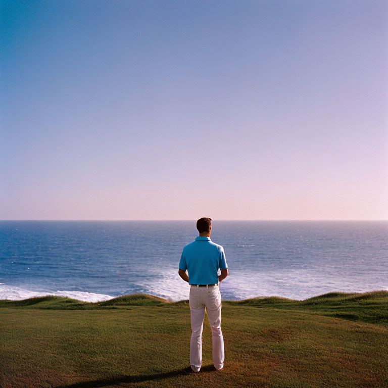Man in blue shirt and white pants on grassy hill by ocean under clear sky