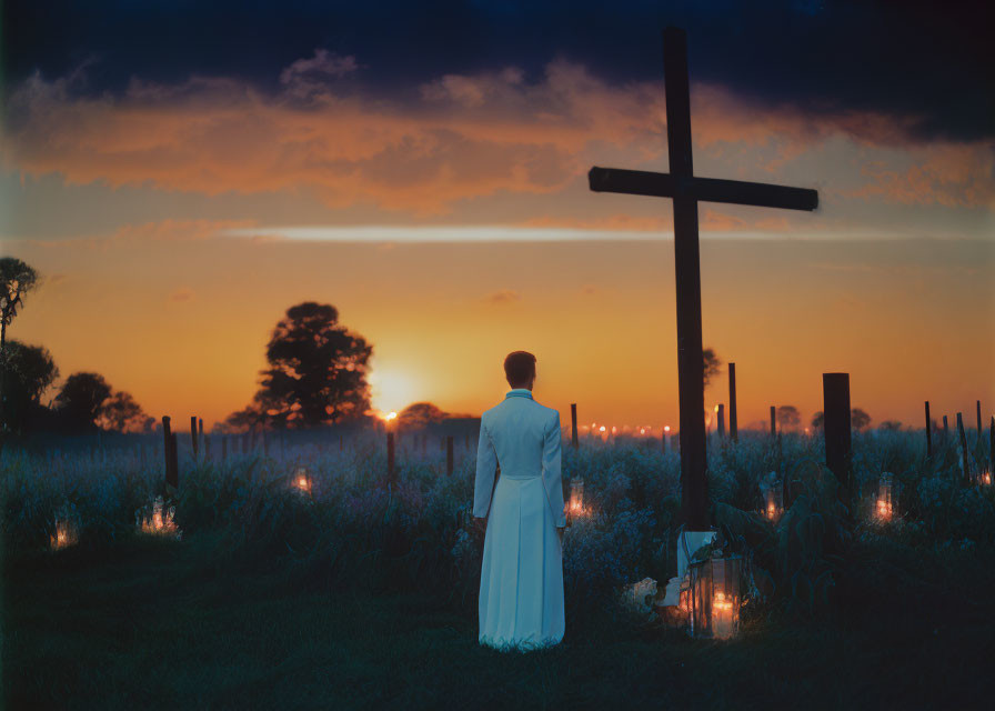 Person in white robe facing sunset with cross silhouette and vibrant sky in rural landscape