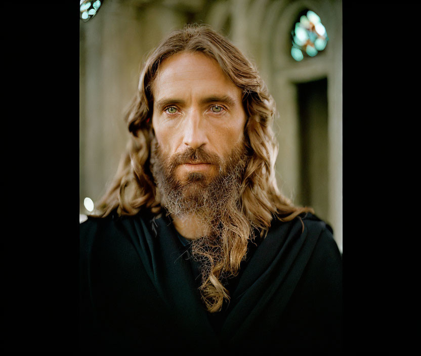 Man with long brown hair and beard in black robe by stained glass windows