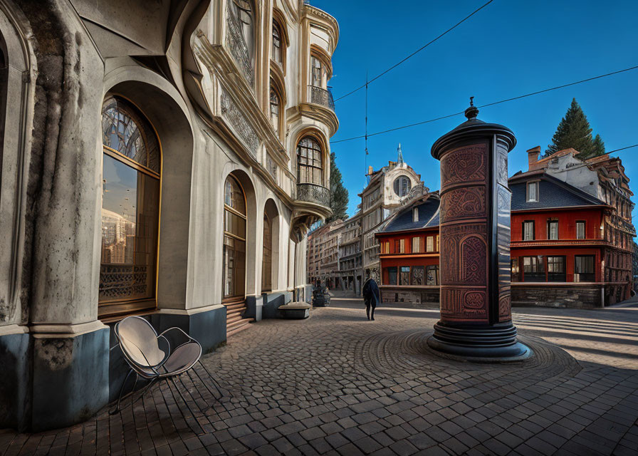 Person walking past red cylindrical kiosk on cobblestone street with ornate buildings and blue sky