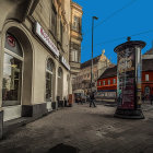 Person walking past red cylindrical kiosk on cobblestone street with ornate buildings and blue sky