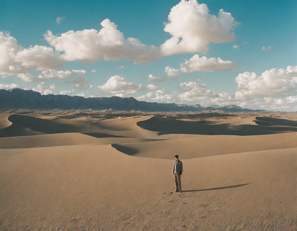 Lonely figure in desert dunes under blue sky and mountains.