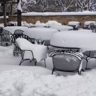 Snow-covered outdoor dining area with white tables and chairs near wooden fence and bushes in snowy ambiance