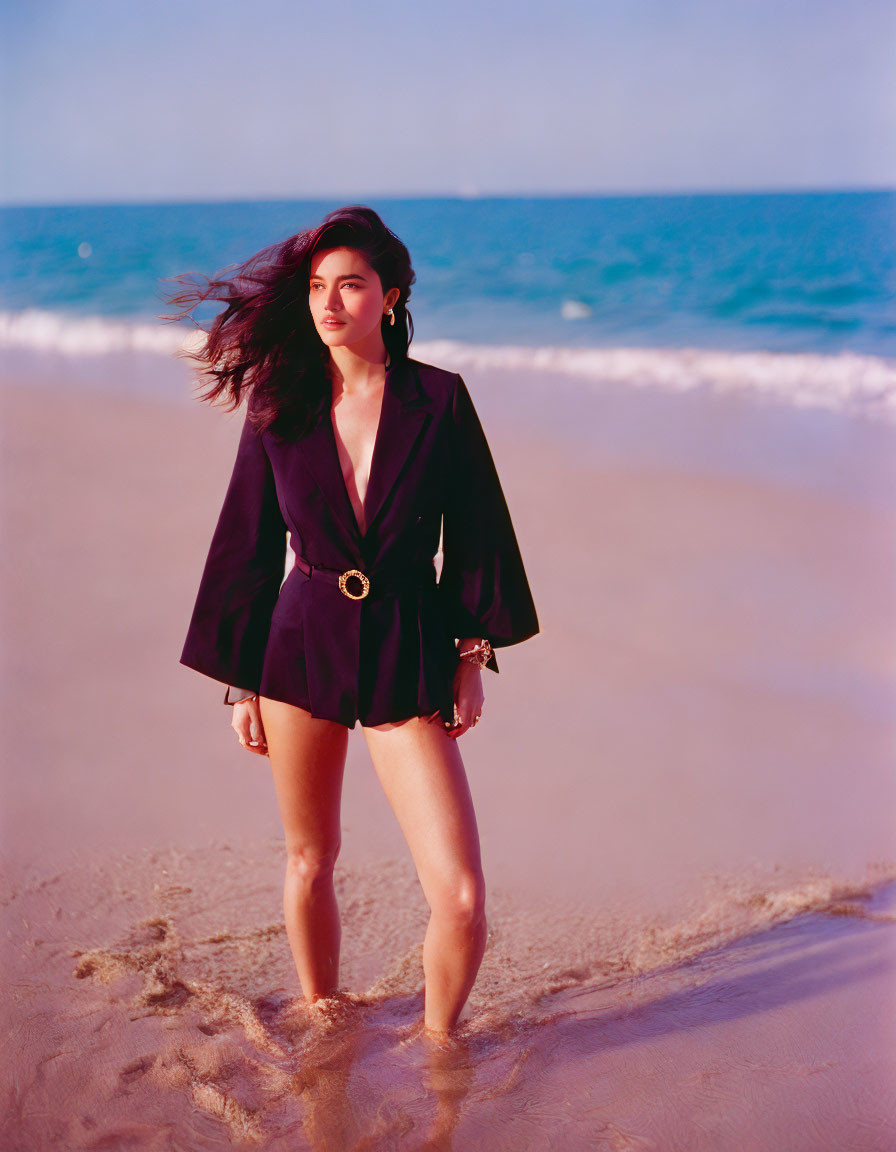 Dark-haired person in black blazer and shorts on sandy beach with clear sky