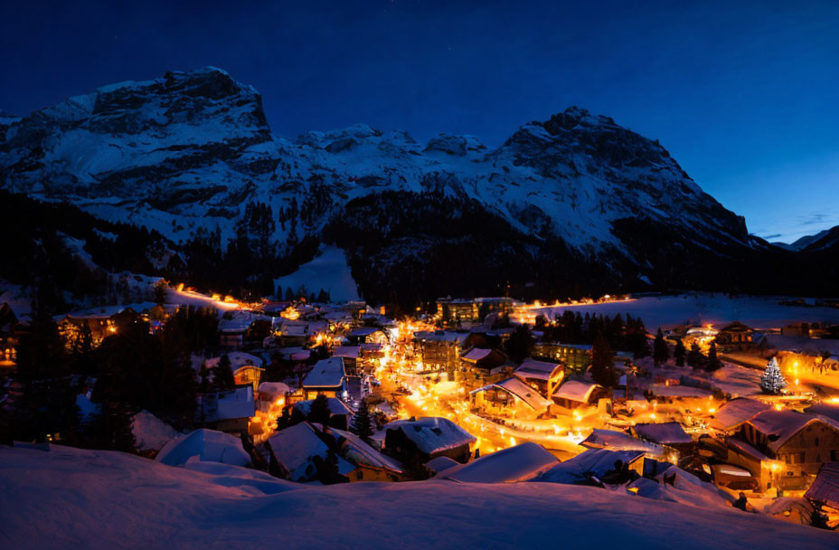 Snow-covered village at twilight nestled by towering mountains under blue sky