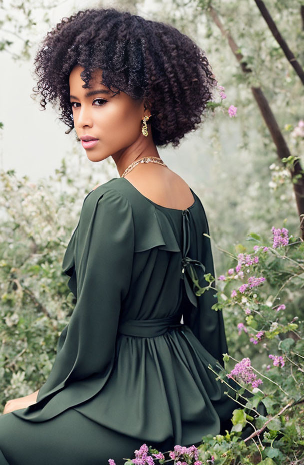 Curly-Haired Woman in Green Dress Sitting Outdoors among Greenery and Purple Flowers